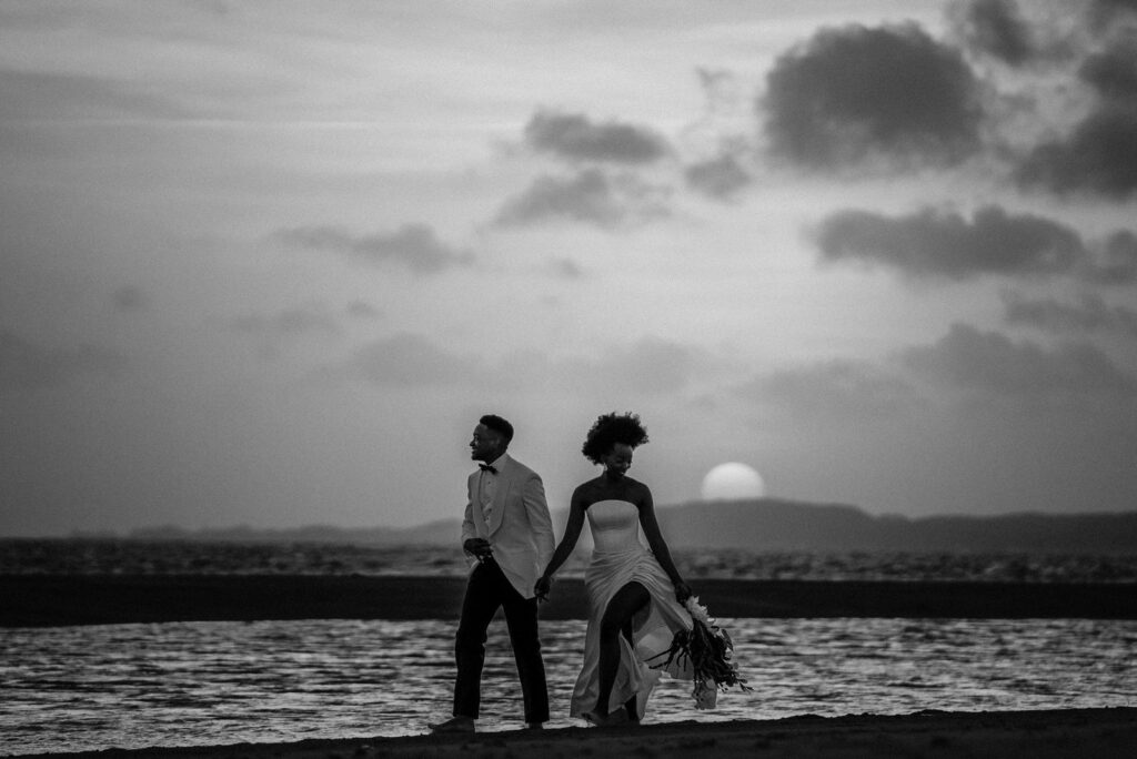 Bride and groom walk along the water as the sun sets in Lamu